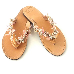 Op de afbeelding: Een paar bruine leren sandalen met roze en witte schelp- en kralenversiering. De sandalen hebben een eenvoudige teenriem en zijn gemaakt in Griekenland.