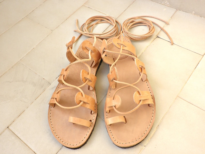 Puede incluir: Un par de sandalias de cuero color canela con cordones que se atan en la pierna. Las sandalias tienen un dise&ntilde;o sencillo con punta redonda y suela plana.