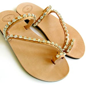 Op de afbeelding: Een paar bruine leren sandalen met een teenring en meerdere banden versierd met heldere strass.