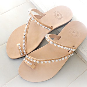 Op de afbeelding: Een paar bruine leren sandalen met strass. De sandalen hebben een eenvoudig ontwerp met een enkele band over de teen en een band om de enkel. De strass zijn in een patroon rond de banden gerangschikt.