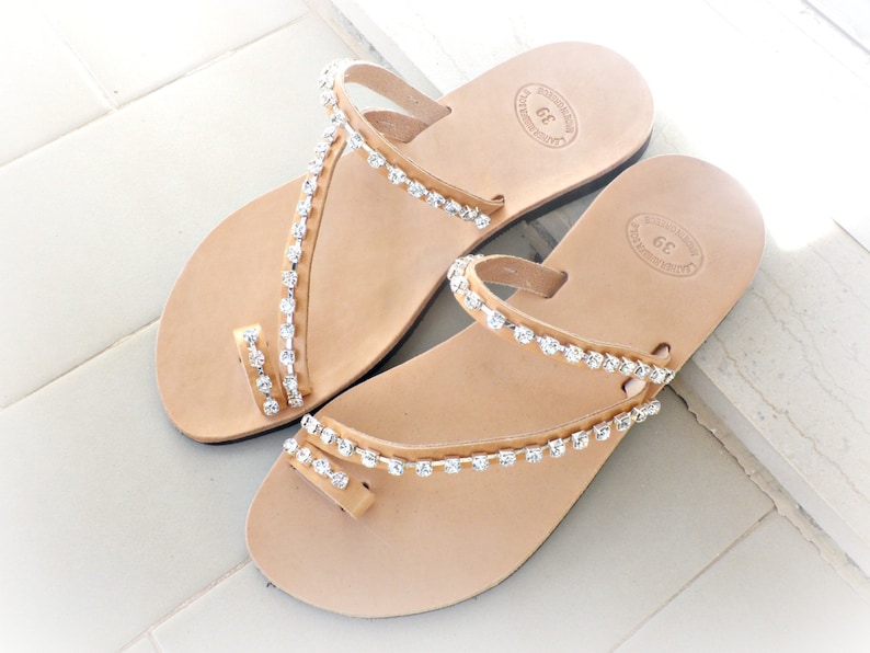Op de afbeelding: Een paar bruine leren sandalen met strass. De sandalen hebben een eenvoudig ontwerp met een enkele band over de teen en een band om de enkel. De strass zijn in een patroon rond de banden gerangschikt.