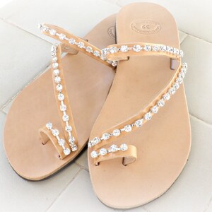 Op de afbeelding: Een paar bruine leren sandalen met een teenriem en meerdere banden versierd met heldere strass.