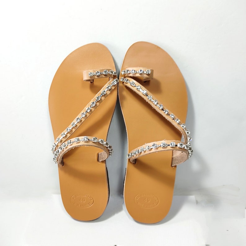 Op de afbeelding: Bruine leren sandalen met twee banden versierd met zilveren strass steentjes. De sandalen hebben een teenlus en een open achterkant.