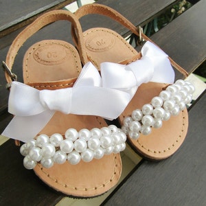 May include: A pair of brown leather sandals with white satin bows and pearl embellishments. The sandals have a simple design and are perfect for a special occasion.
