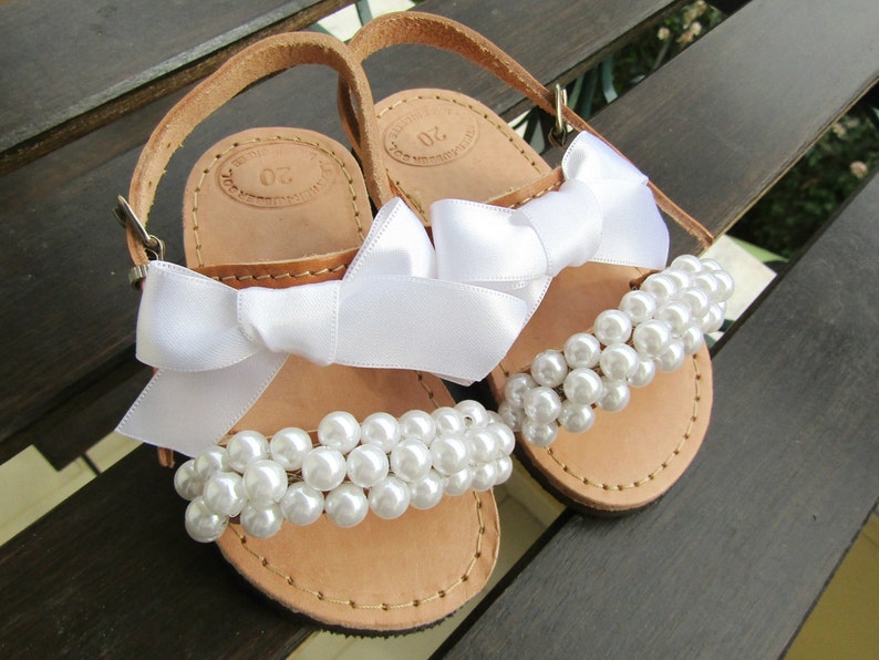 May include: A pair of brown leather sandals with white satin bows and pearl embellishments. The sandals have a simple design and are perfect for a special occasion.