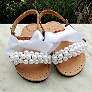 May include: A pair of white leather sandals with white pearl embellishments and white satin bows.