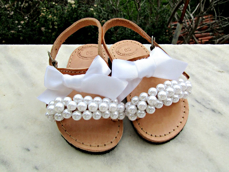 May include: A pair of white leather sandals with white pearl embellishments and white satin bows.