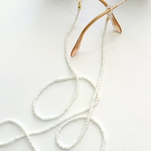 Könnte beinhalten: Weiße Perlen-Brillenschnur mit goldfarbenen Beschlägen. Die Schnur ist um die Bügel einer Sonnenbrille geschlungen.