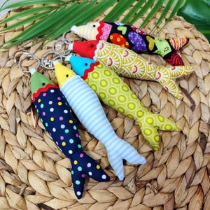 Könnte beinhalten: Eine Sammlung bunter Stoff-Fisch-Schlüsselanhänger. Jeder Fisch hat ein einzigartiges Muster, darunter Punkte, Streifen und florale Designs, mit einem Metall-Schlüsselring. Die Fische sind in Blau-, Grün-, Gelb-, Rot- und Lilatönen gehalten.