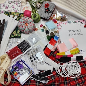 Könnte beinhalten: Eine Sammlung von Nähutensilien, darunter Stoff, Reißverschlüsse, Knöpfe, Bänder, Faden und ein Nähjournal. Die Artikel sind auf einem rot-schwarz karierten Stoff angeordnet. Ein Nadelkissen und eine Schere sind ebenfalls zu sehen.