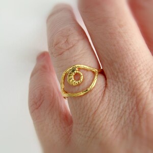 May include: Gold ring with an eye design. The eye is open and has a textured surface. The ring is on a finger.