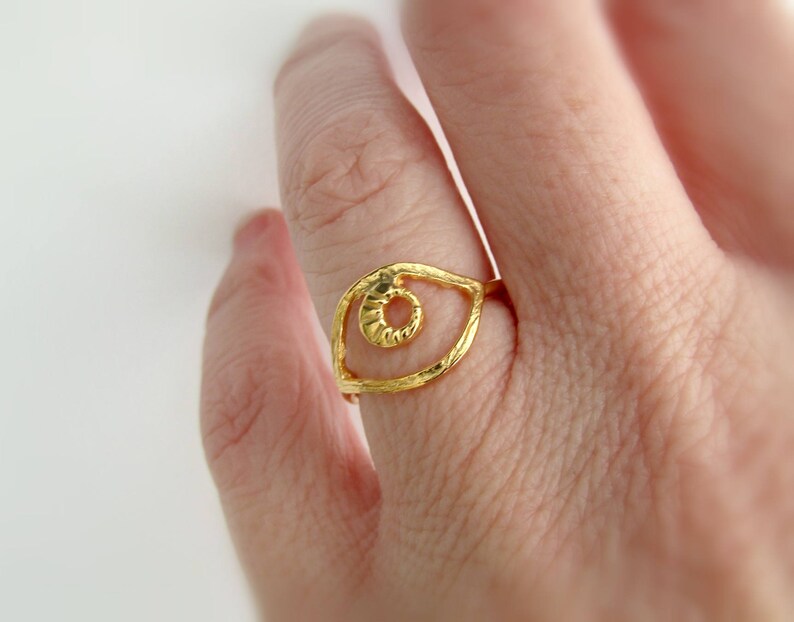 May include: Gold ring with an eye design. The eye is open and has a textured surface. The ring is on a finger.