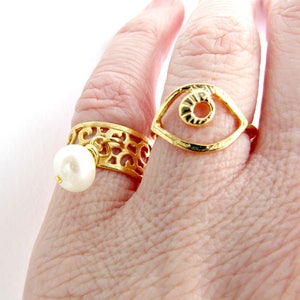 May include: Two gold rings on a finger. One ring has a delicate filigree design with a white pearl dangling from it. The other ring is a simple open circle design with a textured eye shape inside.