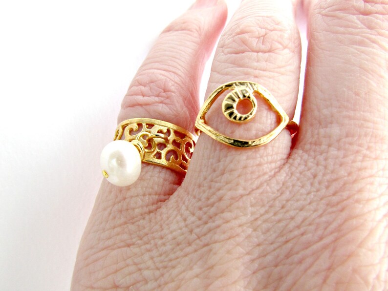 May include: Two gold rings on a finger. One ring has a delicate filigree design with a white pearl dangling from it. The other ring is a simple open circle design with a textured eye shape inside.