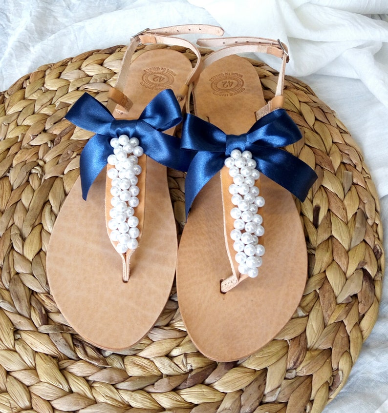 May include: A pair of tan leather sandals with a navy blue ribbon bow and white pearl embellishments.