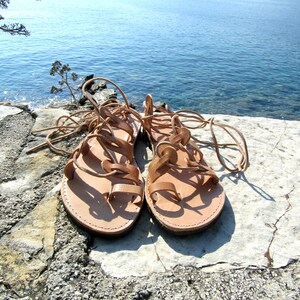 Puede incluir: Un par de sandalias de cuero marr&oacute;n con cordones que se atan en las piernas. Las sandalias est&aacute;n sobre una superficie de piedra gris claro.