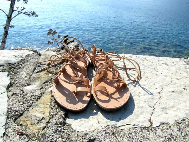 Puede incluir: Un par de sandalias de cuero marr&oacute;n con cordones que se atan en las piernas. Las sandalias est&aacute;n sobre una superficie de piedra gris claro.