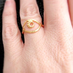 May include: Gold ring with an eye design. The eye is open and has a textured surface. The ring is on a finger.