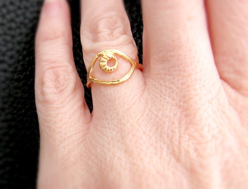 May include: Gold ring with an eye design. The eye is open and has a textured surface. The ring is on a finger.