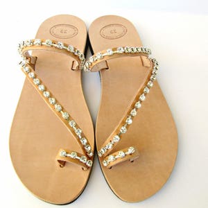 Op de afbeelding: Een paar bruine leren sandalen met een teenring en twee bandjes versierd met heldere strass.