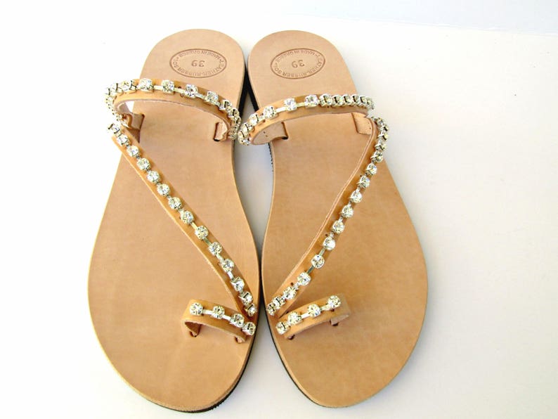 Op de afbeelding: Een paar bruine leren sandalen met een teenring en twee bandjes versierd met heldere strass.