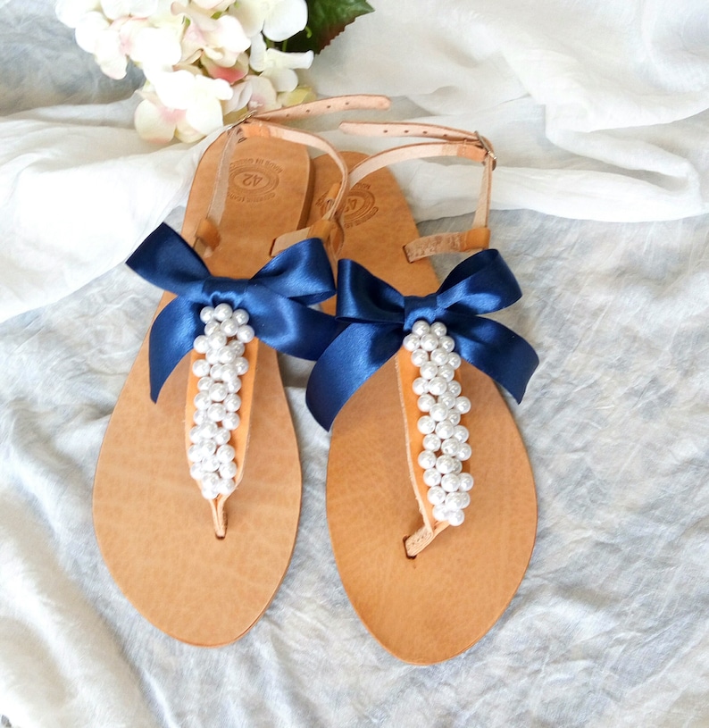 May include: Tan leather sandals with a navy blue satin bow and pearl embellishments. The sandals have a t-strap design and an ankle strap.