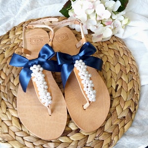 May include: A pair of brown leather sandals with navy blue ribbon bows and white pearl embellishments. The sandals are on a woven straw mat.