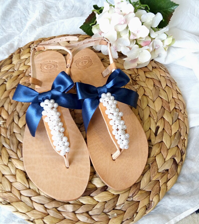 May include: A pair of brown leather sandals with navy blue ribbon bows and white pearl embellishments. The sandals are on a woven straw mat.
