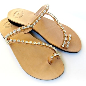 Op de afbeelding: Een paar bruine leren sandalen met een teenring en twee bandjes versierd met zilveren strass.