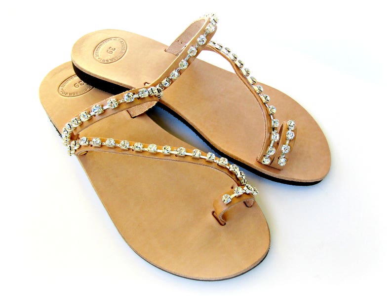 Op de afbeelding: Een paar bruine leren sandalen met een teenring en twee bandjes versierd met zilveren strass.