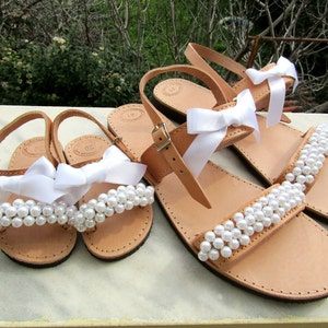 May include: Two pairs of sandals with white pearl embellishments and white satin bows. The sandals are made of brown leather and have a simple design.