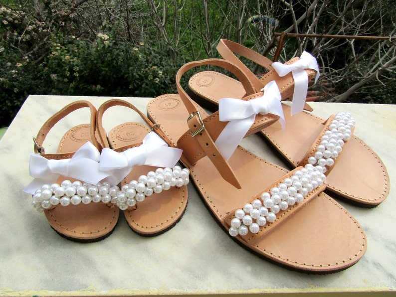 May include: Two pairs of sandals with white pearl embellishments and white satin bows. The sandals are made of brown leather and have a simple design.