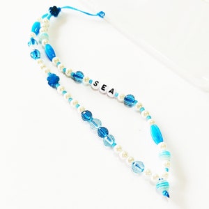 Può includere: Un ciondolo per telefono blu e bianco con la parola "SEA" scritta in perle bianche.