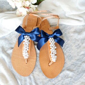 May include: Tan leather sandals with a navy blue ribbon bow and pearl embellishments. The sandals have a t-strap design and an ankle strap.