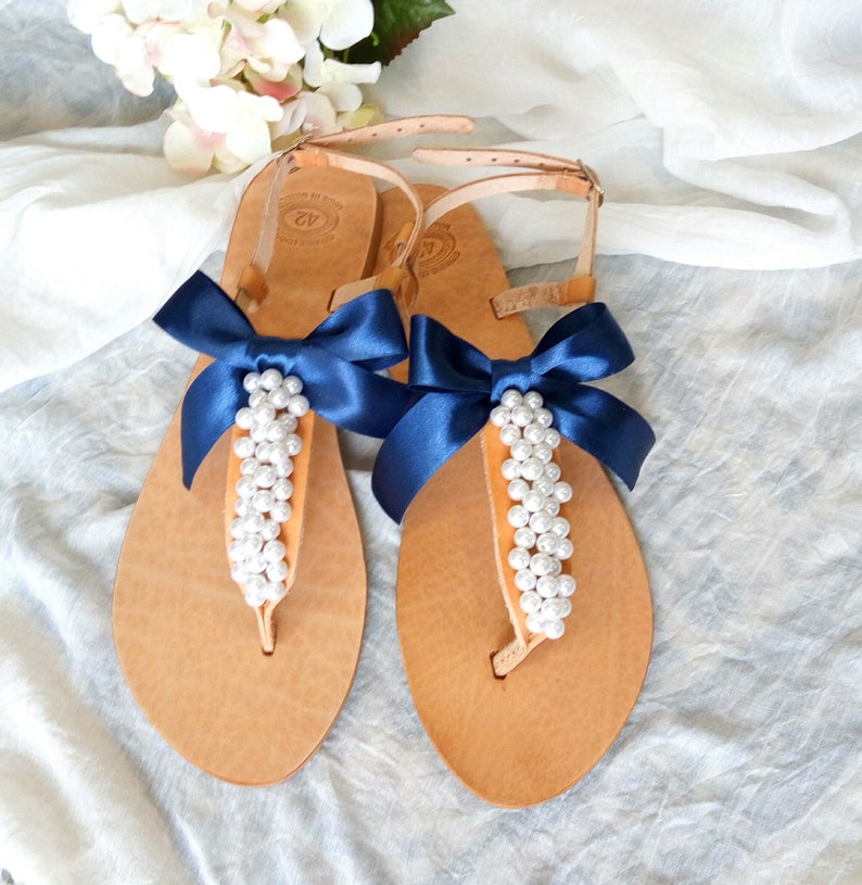 May include: Tan leather sandals with a navy blue ribbon bow and pearl embellishments. The sandals have a t-strap design and an ankle strap.