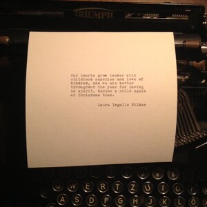 May include: A black and white image of a typewriter with a piece of paper inserted. The paper has a quote from Laura Ingalls Wilder that reads: "Our hearts grow tender with childhood memories and love of kindred, and we are better throughout the year for having in spirit, become a child again at Christmas time."
