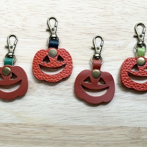 May include: Four pumpkin-shaped keychains made of textured leather in shades of orange and red. Each keychain features a cutout jack-o'-lantern face, a brass-toned metal swivel lobster clasp, and a small colored leather loop secured with a metal rivet.