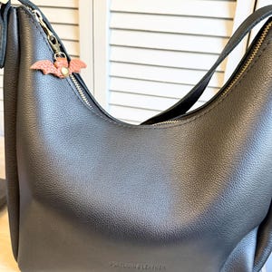May include: Black leather shoulder bag with a gold zipper and a small, pink bat-shaped charm. The bag has a curved design and a strap. The words "PORTLAND & LEATHER" are embossed on the bottom of the bag.