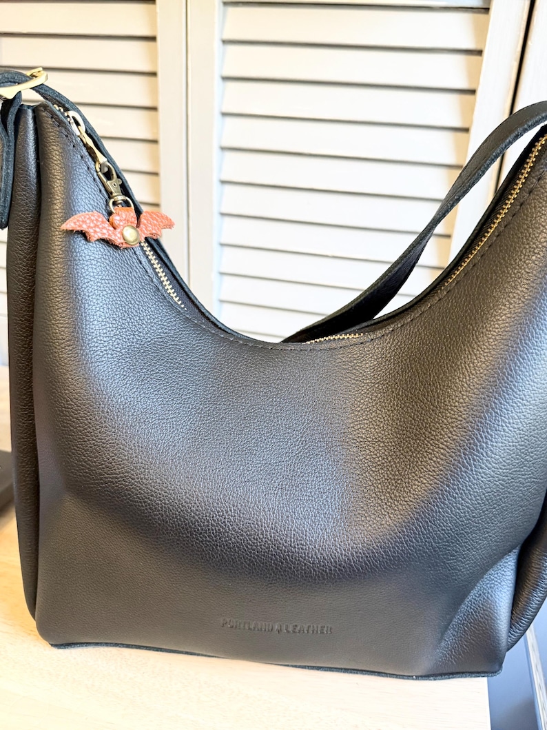 May include: Black leather shoulder bag with a gold zipper and a small, pink bat-shaped charm. The bag has a curved design and a strap. The words "PORTLAND & LEATHER" are embossed on the bottom of the bag.