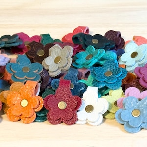 May include: A collection of small, multi-colored leather flower embellishments, each featuring a central brass-toned metal rivet. The flowers are crafted from textured leather in various shades including red, orange, blue, white, and purple.