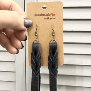 May include: A pair of black leather drop earrings featuring a braided design attached to bronze-toned metal fishhook wires. The earrings are displayed on a rectangular brown card with the text Handmade with love and a red heart.