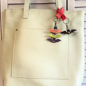 May include: A light green leather tote bag with a front pocket and a pair of bat-shaped keychains. One keychain has three bats in orange, green, and purple. The other keychain has a single black bat.