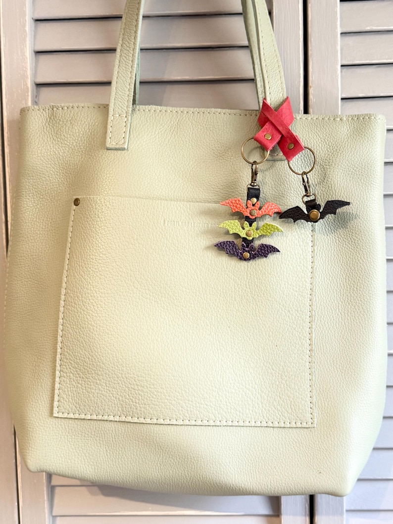 May include: A light green leather tote bag with a front pocket and a pair of bat-shaped keychains. One keychain has three bats in orange, green, and purple. The other keychain has a single black bat.
