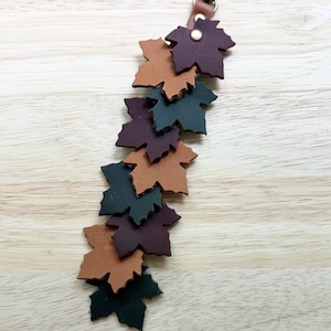 May include: A leather keychain featuring a vertical arrangement of multiple maple leaf-shaped cutouts in shades of dark brown, tan, and forest green. The leaves are attached to a metal swivel lobster clasp with a small brass-colored rivet.