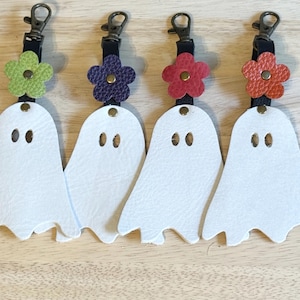 May include: Four white leather ghost keychains with black straps and bronze-colored clasps. Each ghost has two oval eye cutouts and is topped with a flower in green, purple, red, and orange. The keychains are on a light wood surface.