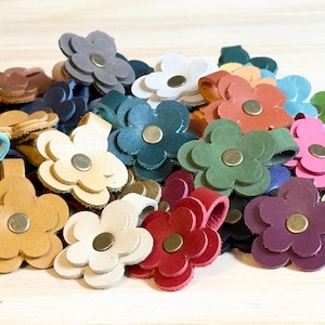 May include: A collection of colorful leather flower-shaped accessories. The flowers come in various colors, including white, red, green, and purple, each with a brass-colored center. These accessories are likely used for crafting or embellishment.
