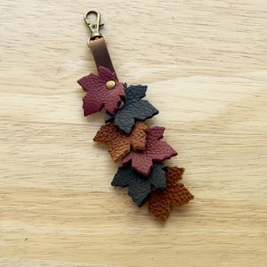 May include: A leather keychain featuring a bronze-toned lobster clasp attached to a brown leather strap. The strap is adorned with multiple layered maple leaf cutouts in textured shades of burgundy, dark gray, and tan, secured by a metal rivet.