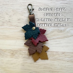 May include: A keychain featuring a metal lobster clasp attached to a brown leather strap with four leaf-shaped cutouts in forest green, chili red, mondelo brandy, and pebbled mango colors, each with a textured surface and a central brass rivet.