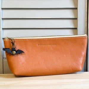 May include: A tan leather pencil case with a zipper closure and a small black bat charm. The text "PORTLAND LEATHER GOODS" is embossed on the case. The case is made of a smooth, textured leather.