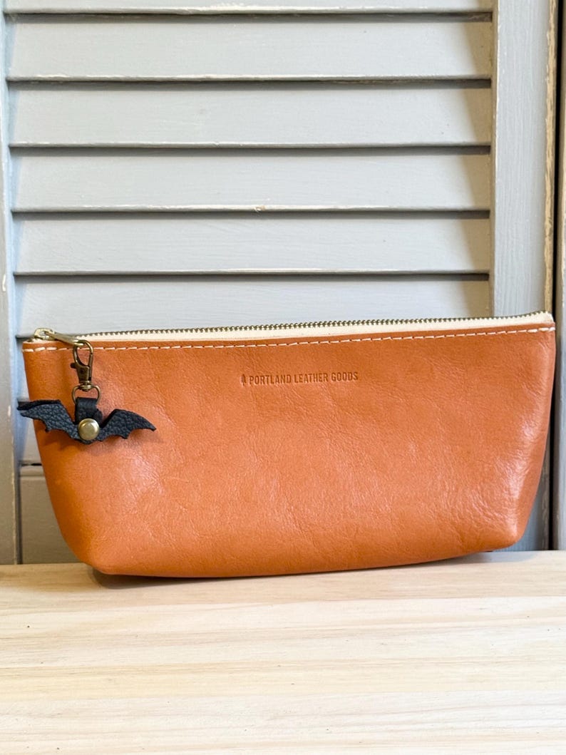 May include: A tan leather pencil case with a zipper closure and a small black bat charm. The text "PORTLAND LEATHER GOODS" is embossed on the case. The case is made of a smooth, textured leather.